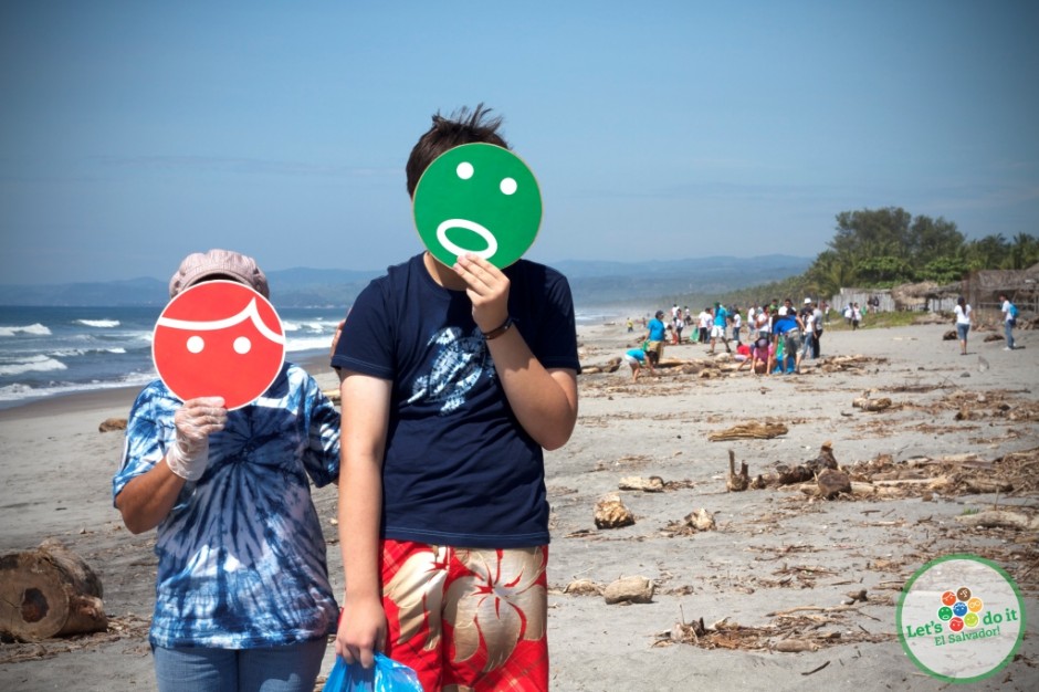 two children on a beach holding a green and red cardboard smiley face in front of their faces