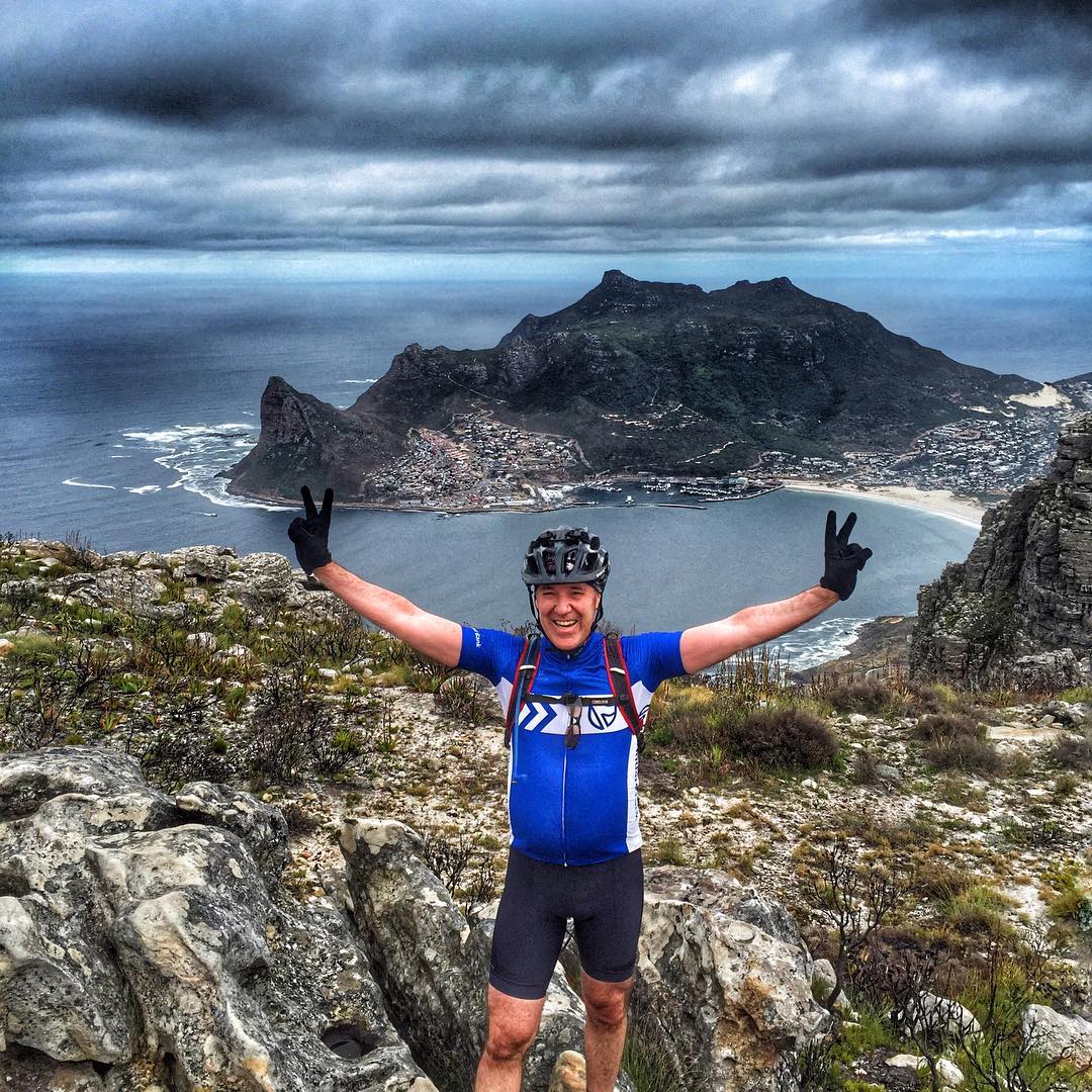 man in blue cycling gear standing above Noordhoek Peak, arms outstretched